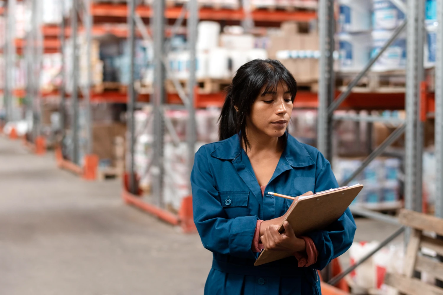 Woman holding a clipboard while inspecting items in a warehouse aisle.